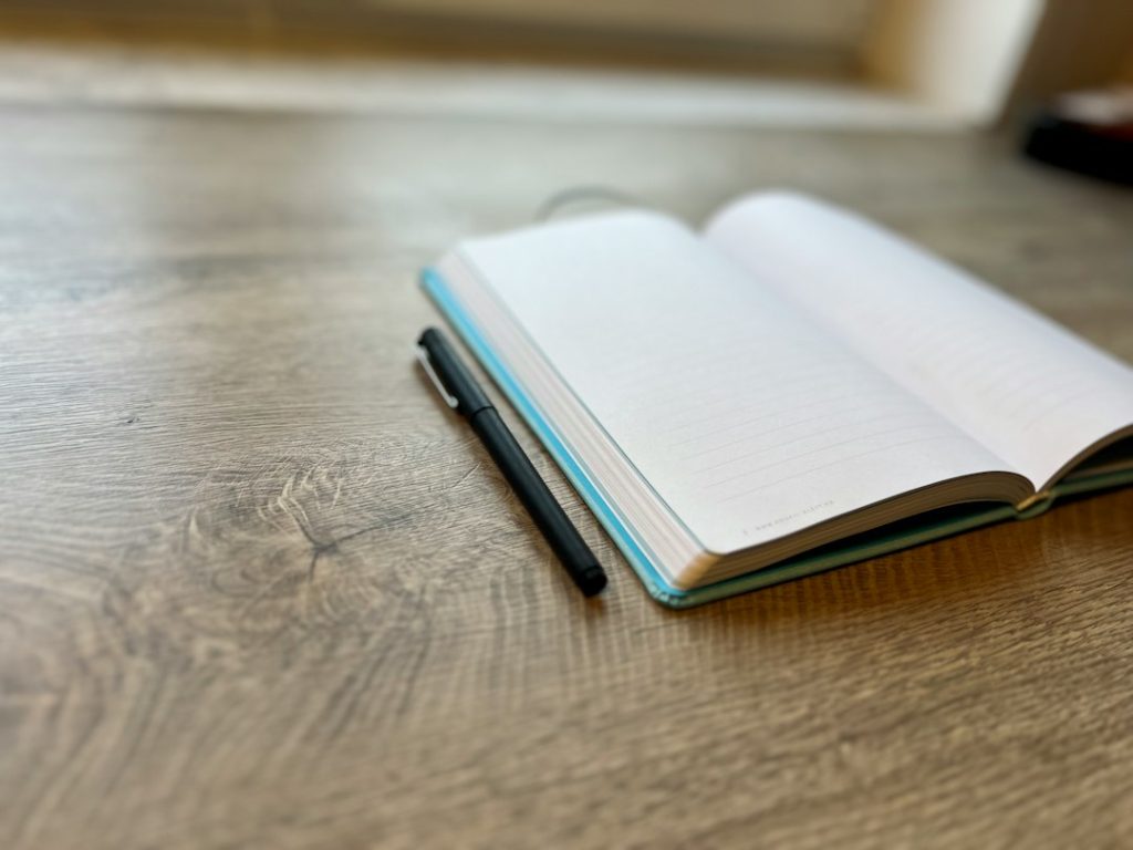 An open blank notebook with a black pen beside it on a wooden desk