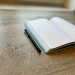 An open blank notebook with a black pen beside it on a wooden desk