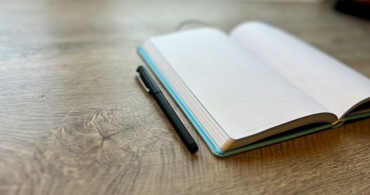 An open blank notebook with a black pen beside it on a wooden desk