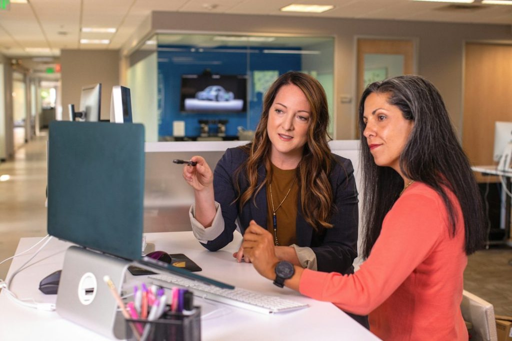 Two women in an office sitting at a desk, one pointing a pen toward a laptop while the other listens