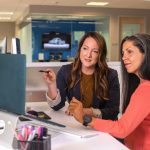 Two women in an office sitting at a desk, one pointing a pen toward a laptop while the other listens