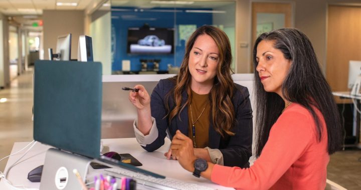 Two women in an office sitting at a desk, one pointing a pen toward a laptop while the other listens