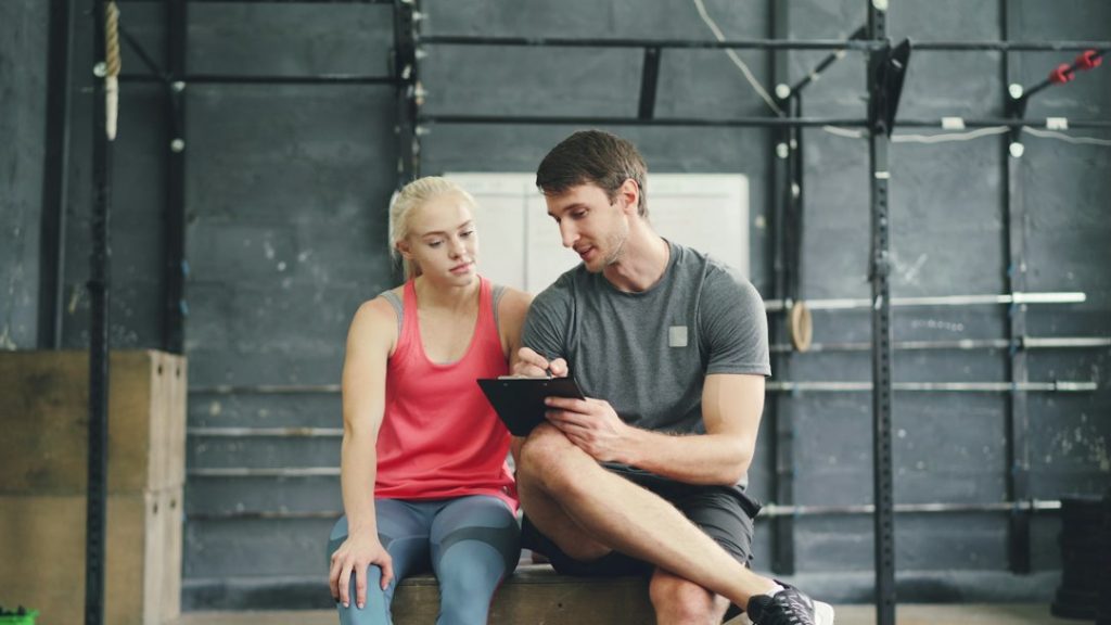 A man in a gray t-shirt sitting on a gym box writes on a clipboard while a woman in a pink tank top beside him looks on, in a one-on-one coaching conversation