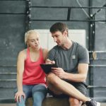 A man in a gray t-shirt sitting on a gym box writes on a clipboard while a woman in a pink tank top beside him looks on, in a one-on-one coaching conversation