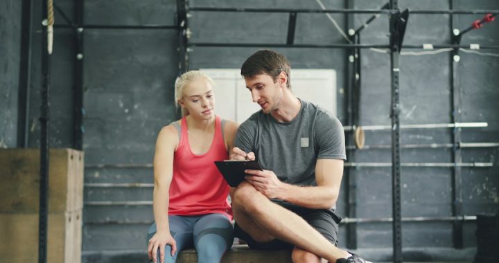 A man in a gray t-shirt sitting on a gym box writes on a clipboard while a woman in a pink tank top beside him looks on, in a one-on-one coaching conversation